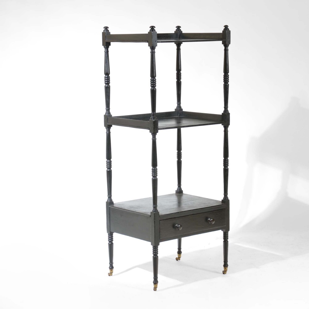 A three-tiered Georgian ebonised wood whatnot featuring turned support columns, top corner finials, a lower drawer with twin knob handles, and brass castors.