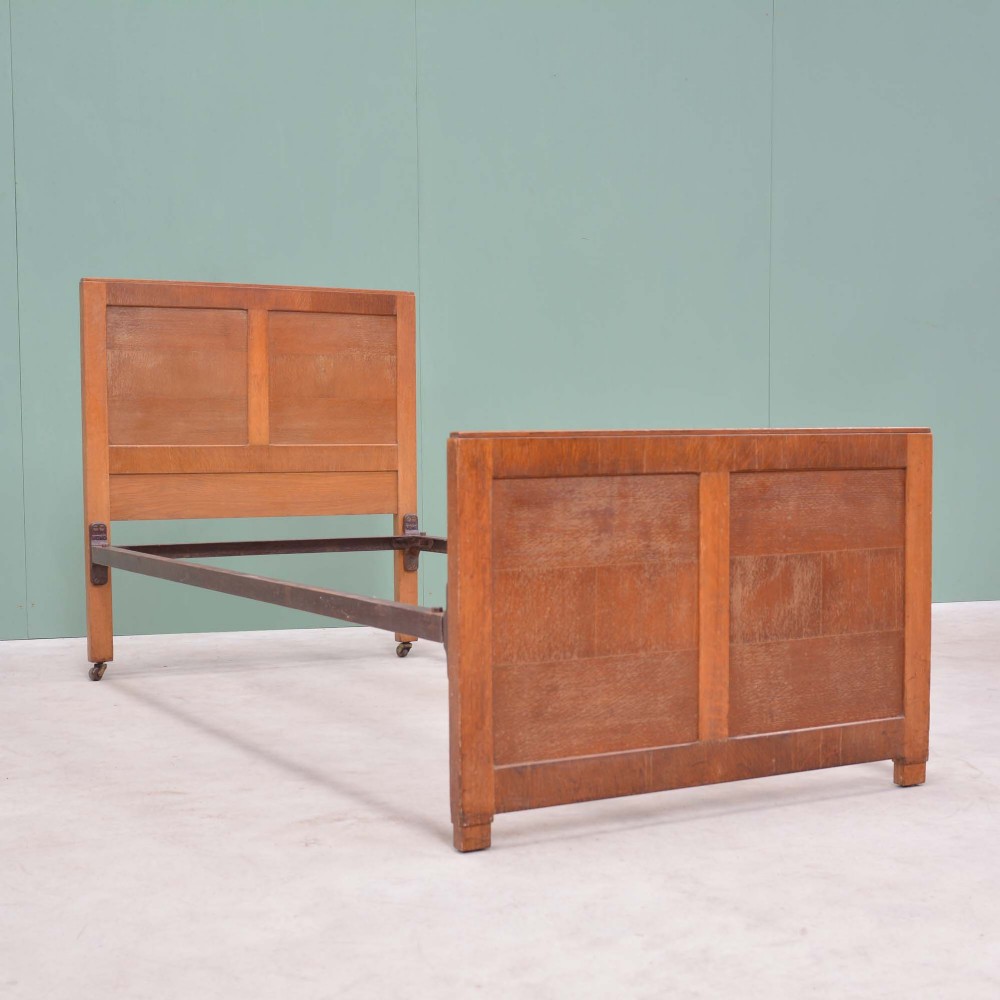 An oak single bed frame featuring a simple panelled headboard and footboard connected by iron side rails on small castors.