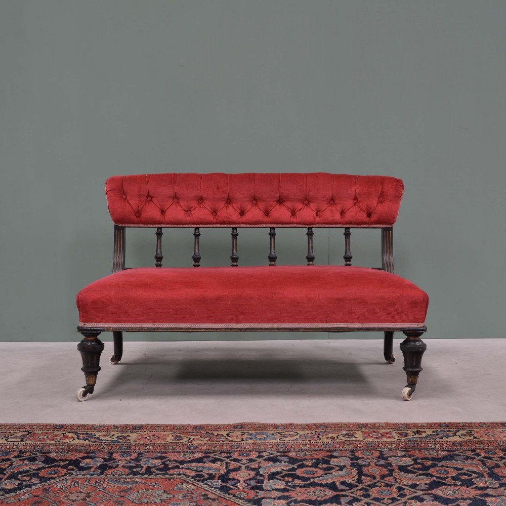 An Aesthetic Movement ebonised wooden settee featuring red velvet button-back upholstery, turned spindle supports, and turned legs on ceramic castors.