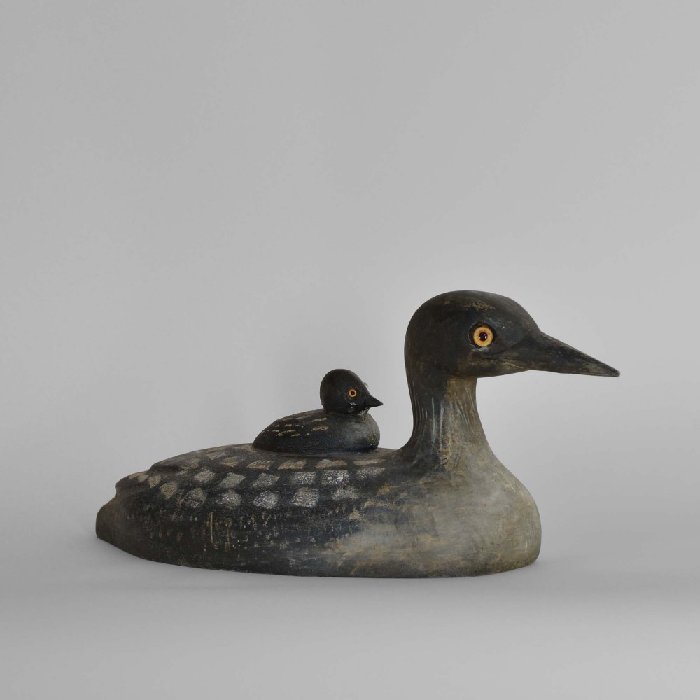 A hand-carved wooden loon decoy painted in dark grey with white spotted patterns, featuring a smaller loon chick sculpted on its back.