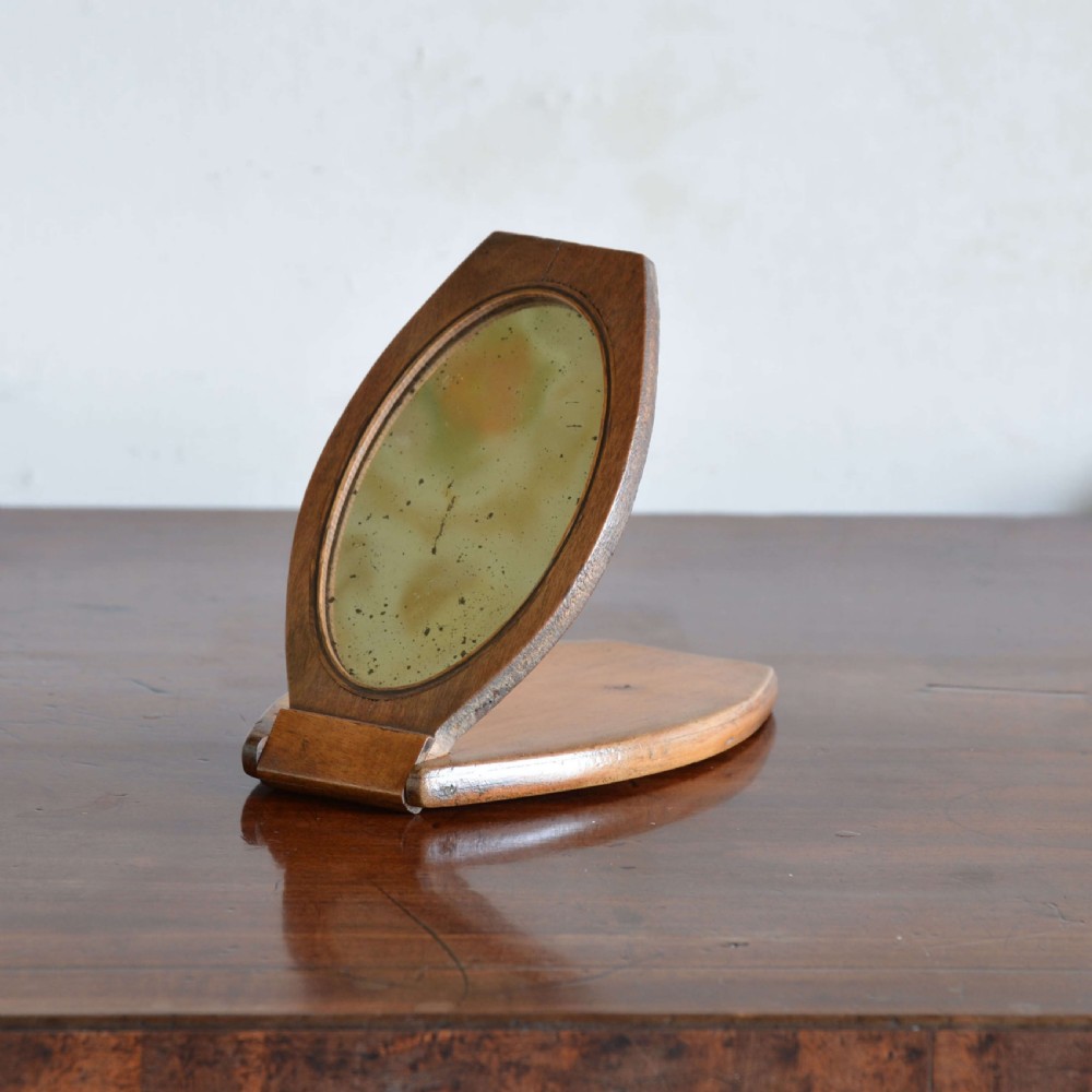 A hinged walnut campaign shaving mirror with an oval glass plate resting open on a wooden surface.