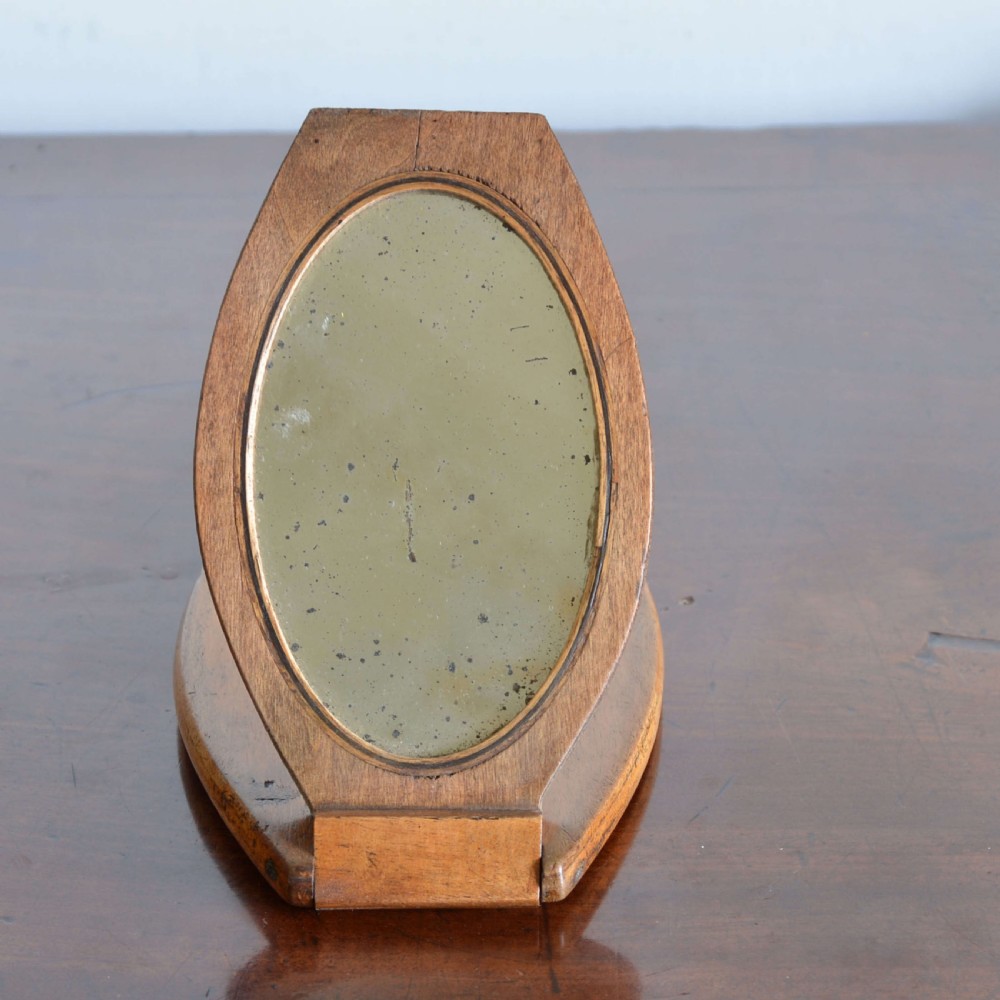 A hinged walnut campaign shaving mirror with an oval glass plate resting open on a wooden surface. View V