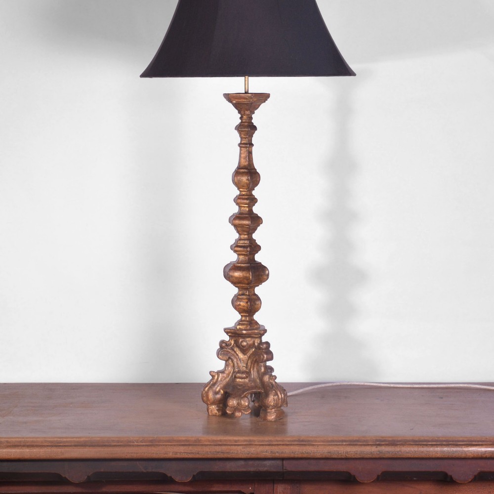 An 18th-century carved giltwood altar candlestick converted into a tall table lamp with a flared dark lampshade.