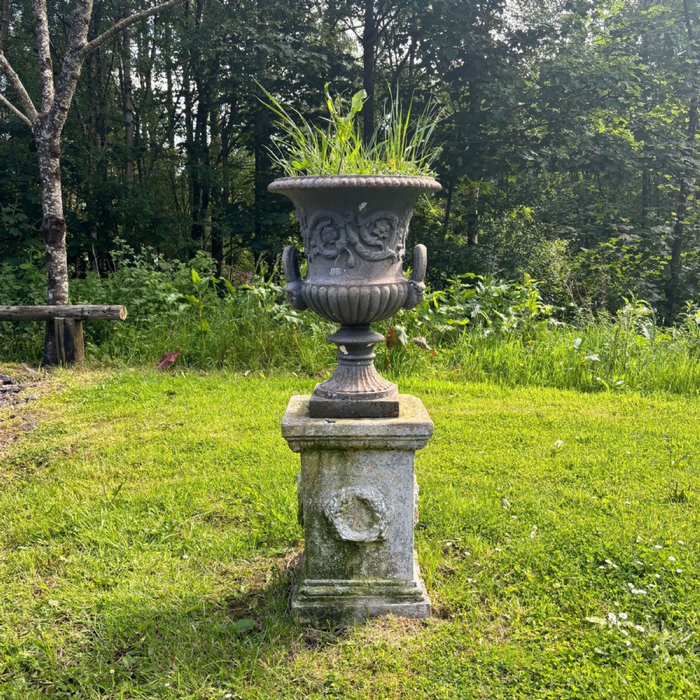 A Victorian bell-shaped cast iron campana urn featuring relief carving and loop handles set upon a weathered square stone pedestal.