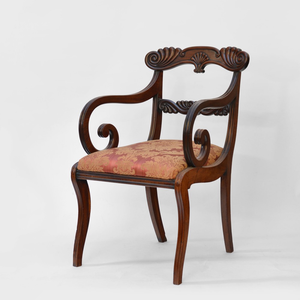 A Regency mahogany armchair featuring carved scroll arms, curved sabre legs, an ornate back rail, and gold patterned upholstery.