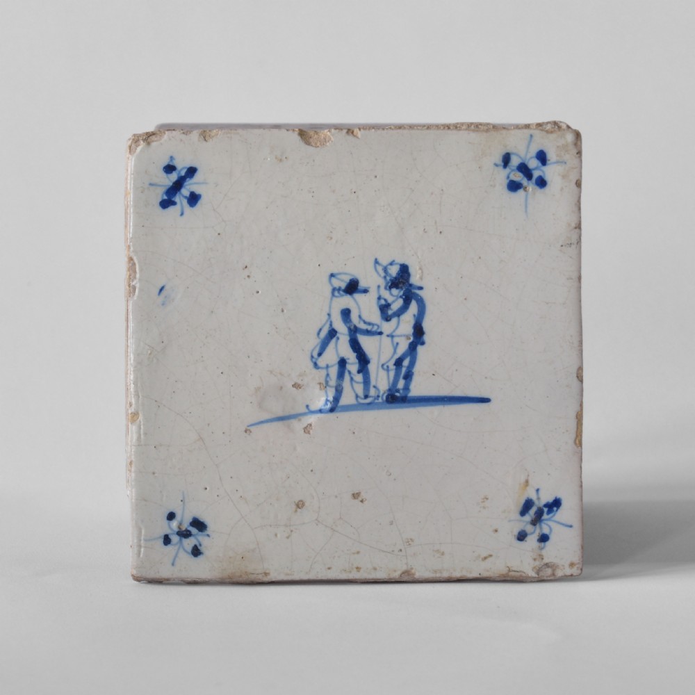 A square 18th-century tin-glazed earthenware tile decorated with hand-painted blue figures in the centre and small corner motifs against an off-white background. View II