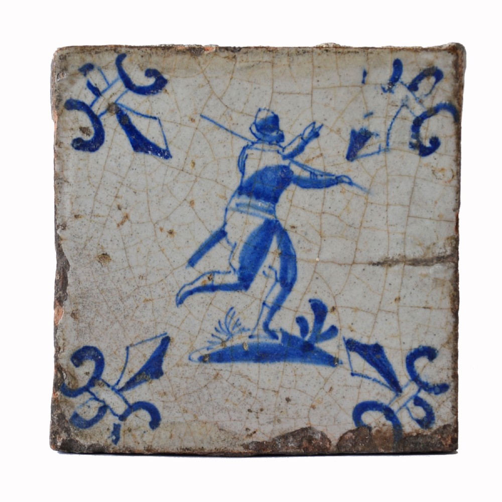 A square 17th century tin-glazed earthenware tile featuring a blue hand-painted soldier with a spear, surrounded by corner motifs on a crazed white glaze background. View II