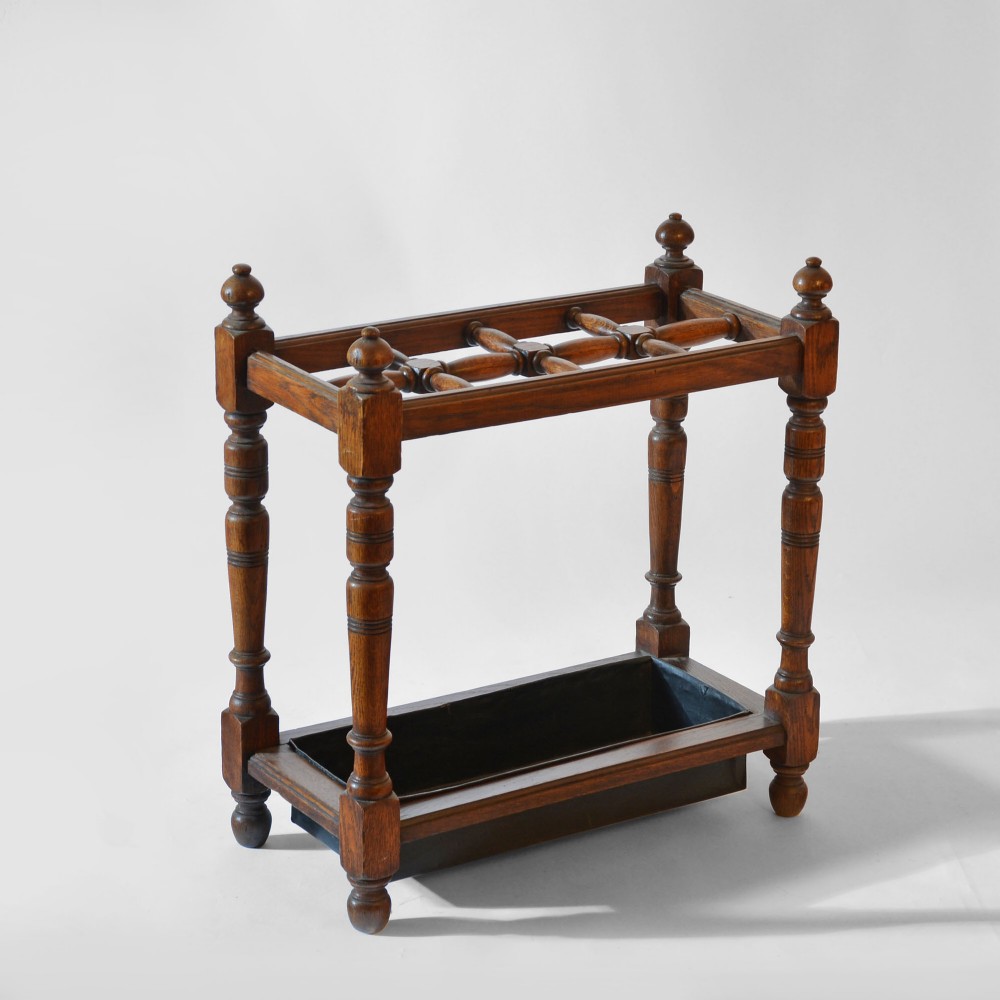An antique dark oak umbrella stand featuring turned upright legs, ball finials, partitioned top dividers, and a metal drip tray at the base.