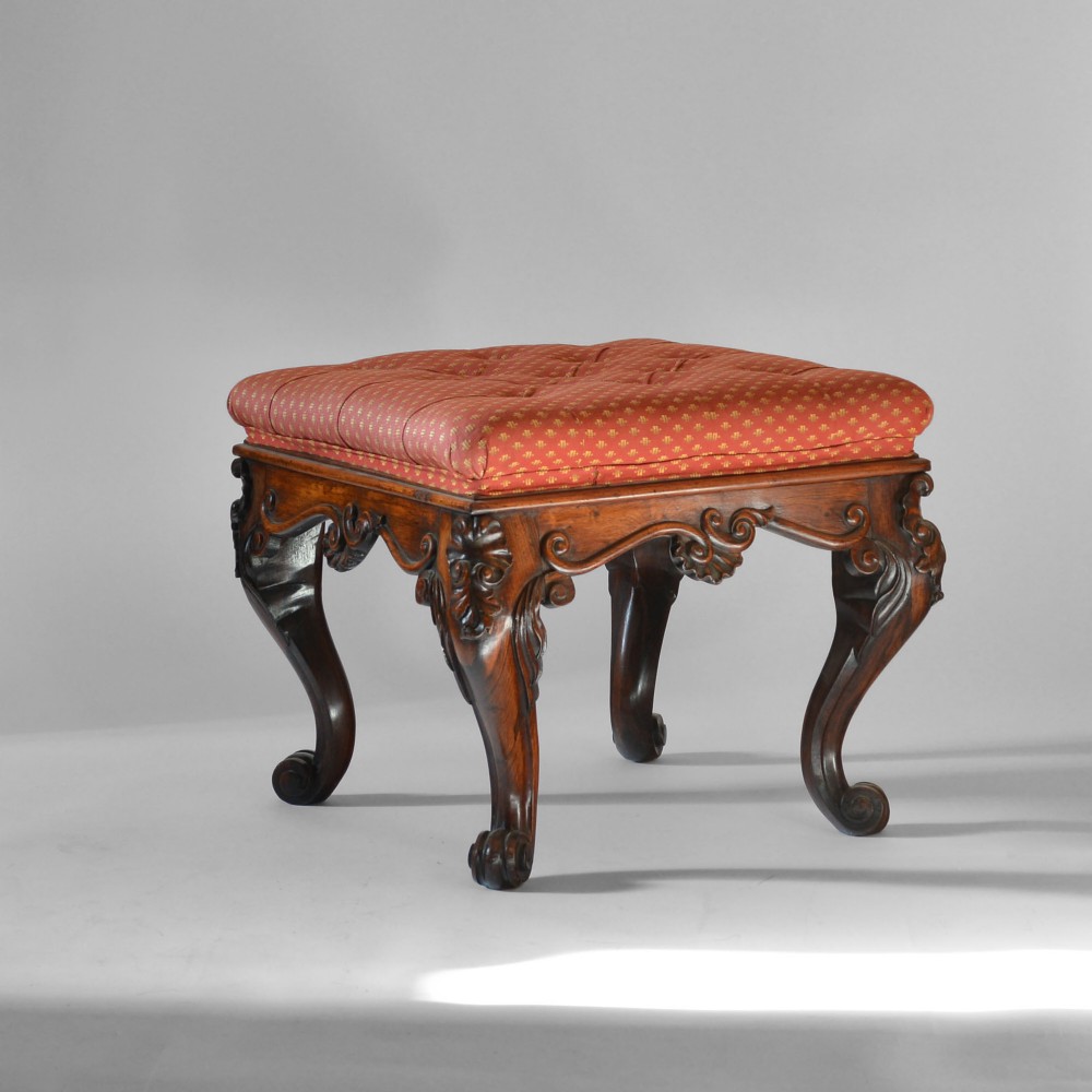 A rectangular Anglo-Indian rosewood footstool featuring a red upholstered buttoned seat above deeply carved aprons and cabriole legs with scroll feet.