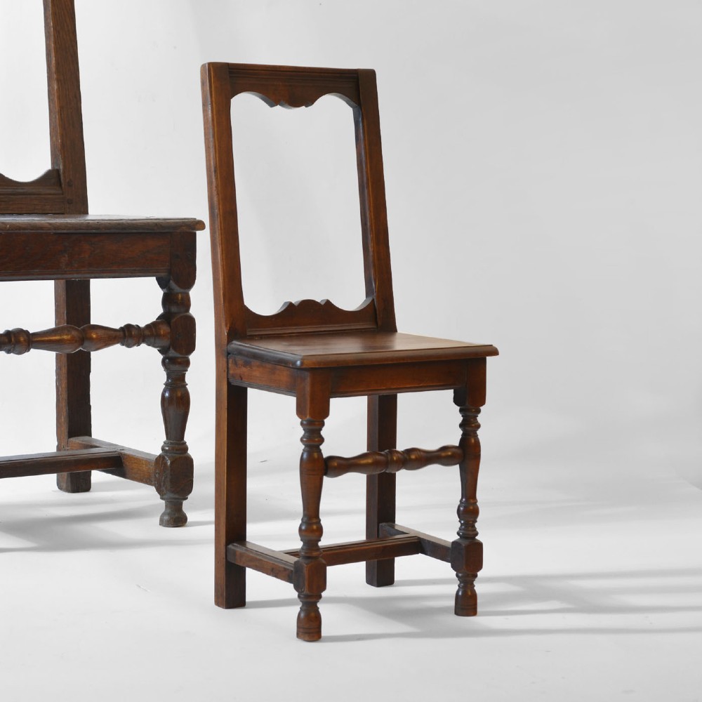 A small, warm brown antique fruitwood child chair featuring an open rectangular back with contoured cut-outs, a solid seat, and turned front legs with stretchers.