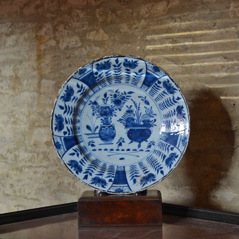 A circular tin-glazed earthenware charger featuring hand-painted cobalt blue urn and botanical motifs on a white background with a panelled rim.