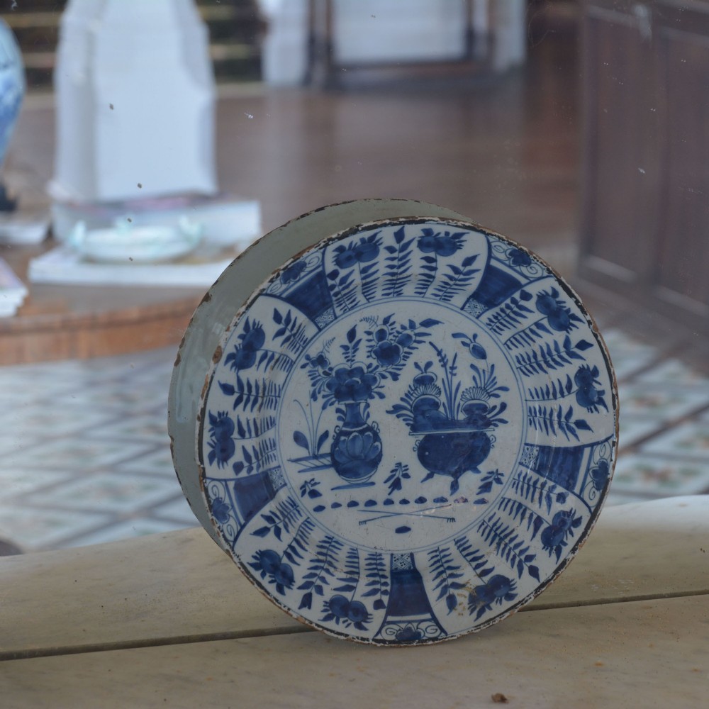 A circular tin-glazed earthenware charger featuring hand-painted cobalt blue urn and botanical motifs on a white background with a panelled rim. View V