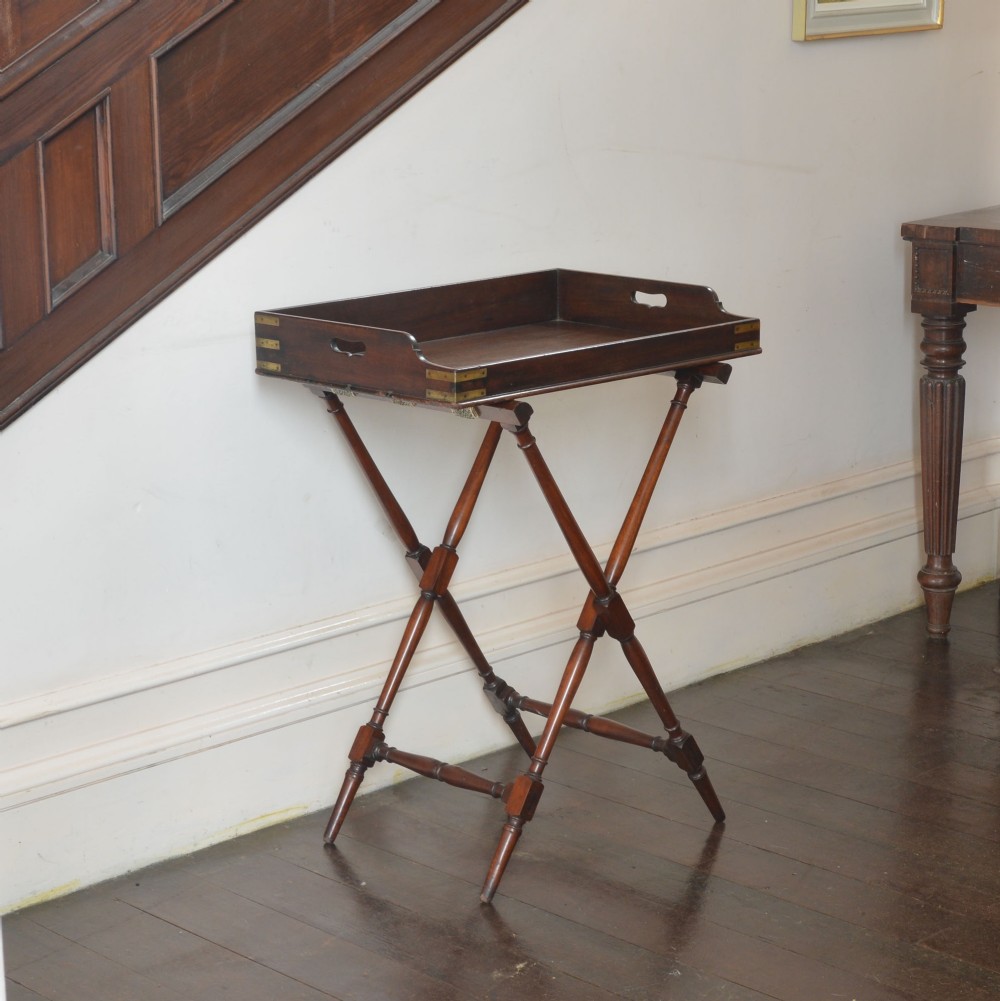 An antique dark mahogany butler tray with gallery edges and brass corner brackets set atop a folding x-frame stand with turned wooden legs.