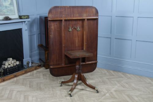 fig 6. A Regency mahogany tilt-top dining table featuring a rectangular crossbanded top supported by a turned pedestal column and four reeded sabre legs with brass castors.