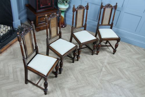 fig 1. A set of four dark oak Jacobean Revival dining chairs featuring woven cane backs, carved crests, turned legs, and cream fabric seats.