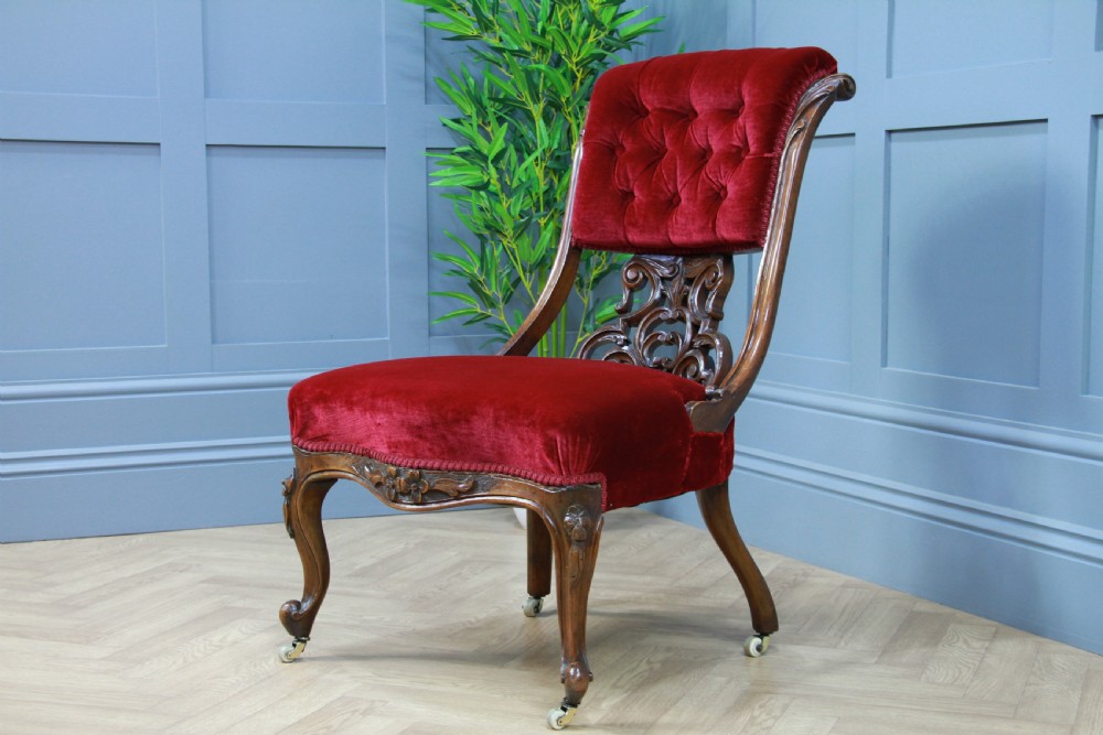 A Victorian carved walnut nursing chair featuring deep crimson red velvet buttoned upholstery, openwork fretwork backrest detail, and cabriole front legs on castors. View II