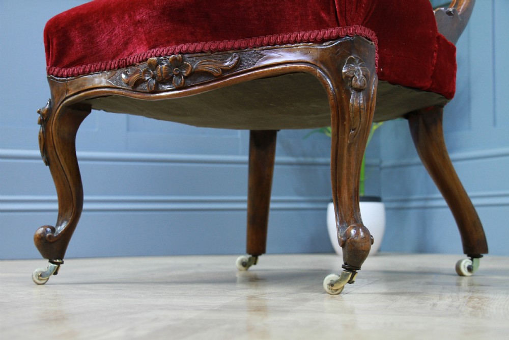 A Victorian carved walnut nursing chair featuring deep crimson red velvet buttoned upholstery, openwork fretwork backrest detail, and cabriole front legs on castors. View III