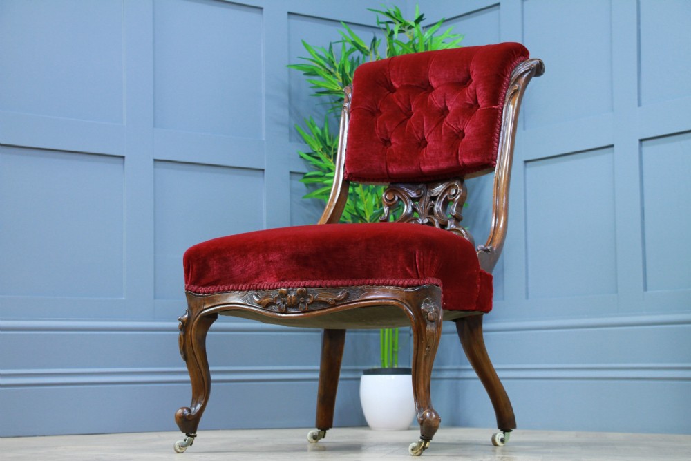 A Victorian carved walnut nursing chair featuring deep crimson red velvet buttoned upholstery, openwork fretwork backrest detail, and cabriole front legs on castors. View IV