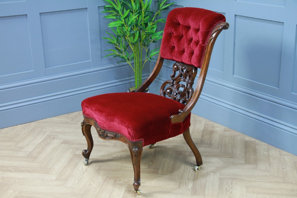 A Victorian carved walnut nursing chair featuring deep crimson red velvet buttoned upholstery, openwork fretwork backrest detail, and cabriole front legs on castors. View V
