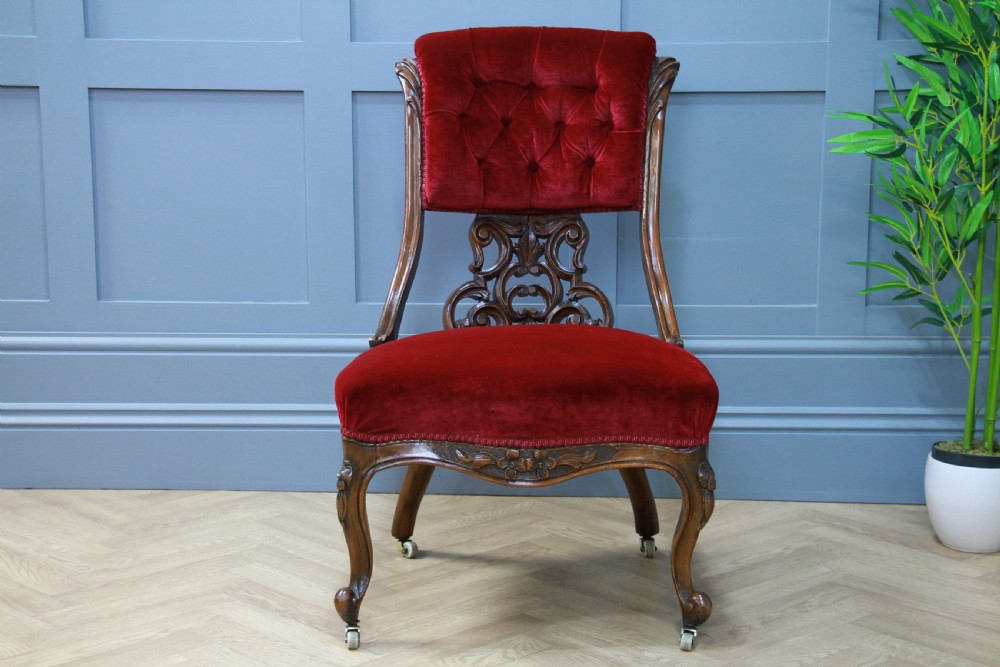 A Victorian carved walnut nursing chair featuring deep crimson red velvet buttoned upholstery, openwork fretwork backrest detail, and cabriole front legs on castors. View VI
