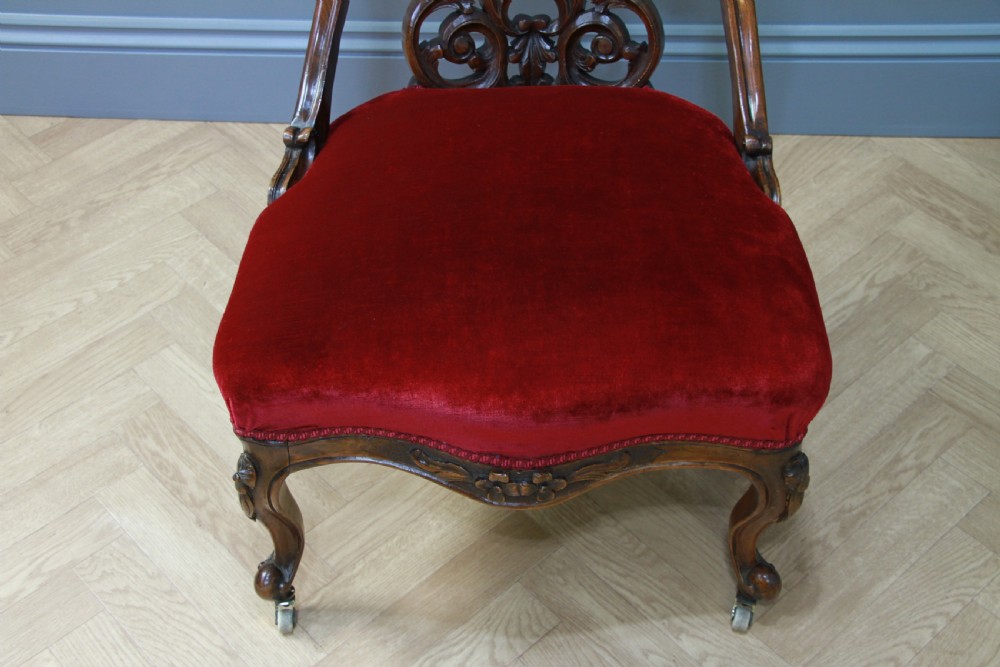 A Victorian carved walnut nursing chair featuring deep crimson red velvet buttoned upholstery, openwork fretwork backrest detail, and cabriole front legs on castors. View VII