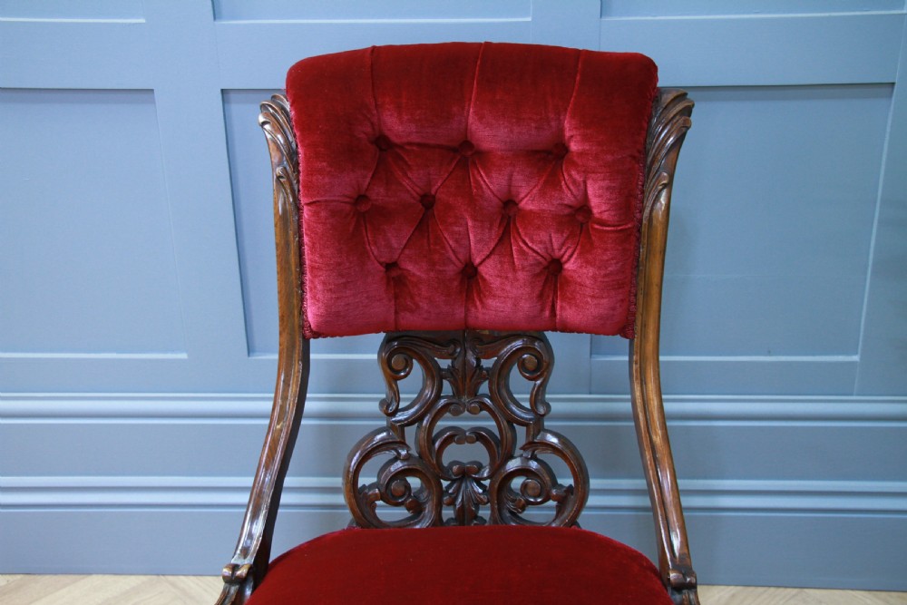 A Victorian carved walnut nursing chair featuring deep crimson red velvet buttoned upholstery, openwork fretwork backrest detail, and cabriole front legs on castors. View VIII