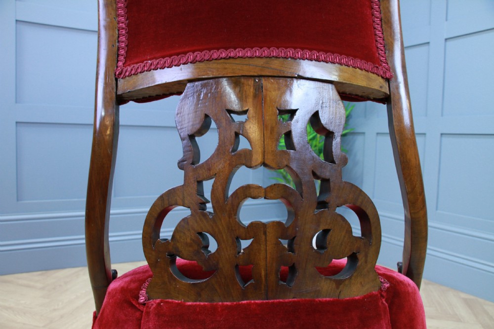 A Victorian carved walnut nursing chair featuring deep crimson red velvet buttoned upholstery, openwork fretwork backrest detail, and cabriole front legs on castors. View IX