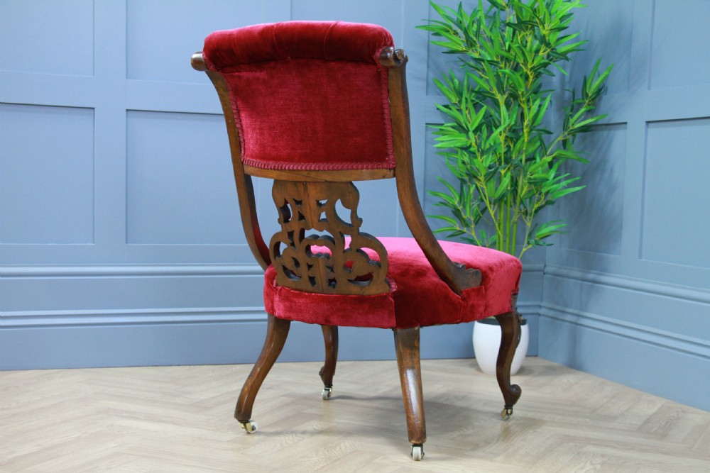 A Victorian carved walnut nursing chair featuring deep crimson red velvet buttoned upholstery, openwork fretwork backrest detail, and cabriole front legs on castors. View X