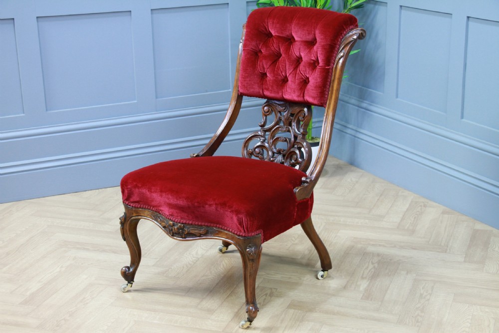 A Victorian carved walnut nursing chair featuring deep crimson red velvet buttoned upholstery, openwork fretwork backrest detail, and cabriole front legs on castors. View XI