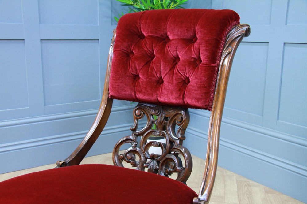 A Victorian carved walnut nursing chair featuring deep crimson red velvet buttoned upholstery, openwork fretwork backrest detail, and cabriole front legs on castors. View XII