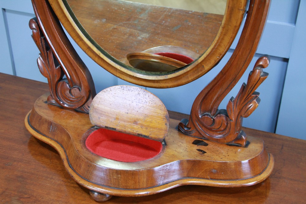 A Victorian mahogany swing toilet mirror featuring an oval moulded frame, carved scroll upright supports, and a shaped plinth base. View V