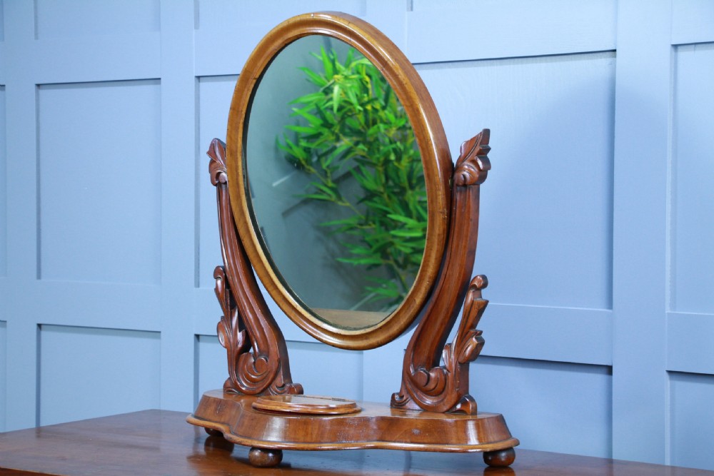 A Victorian mahogany swing toilet mirror featuring an oval moulded frame, carved scroll upright supports, and a shaped plinth base. View VI