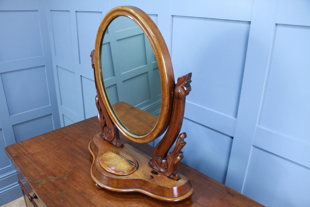 A Victorian mahogany swing toilet mirror featuring an oval moulded frame, carved scroll upright supports, and a shaped plinth base. View X