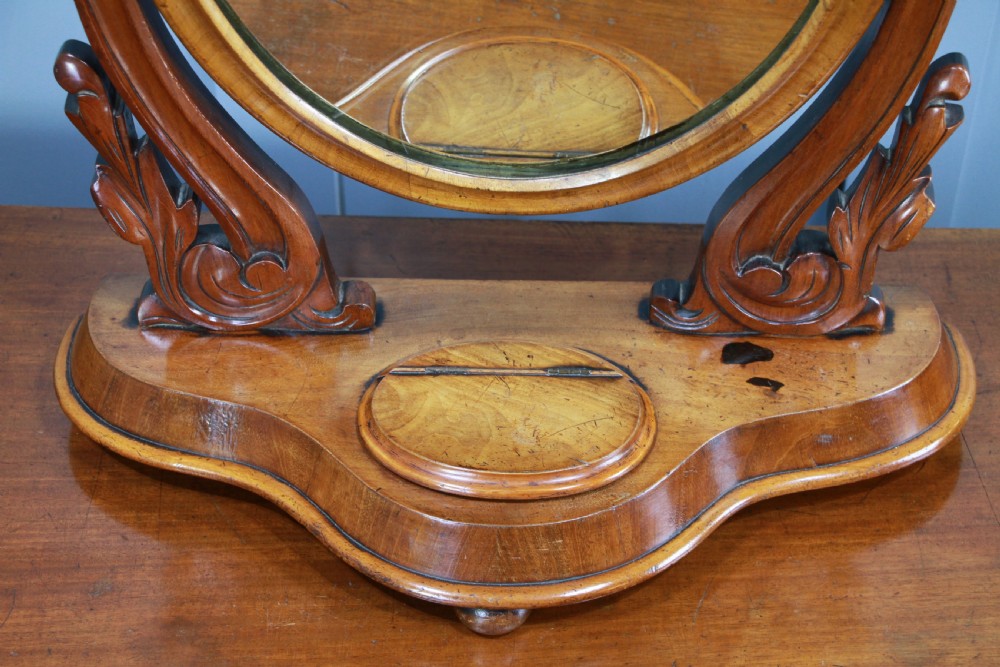 A Victorian mahogany swing toilet mirror featuring an oval moulded frame, carved scroll upright supports, and a shaped plinth base. View XII