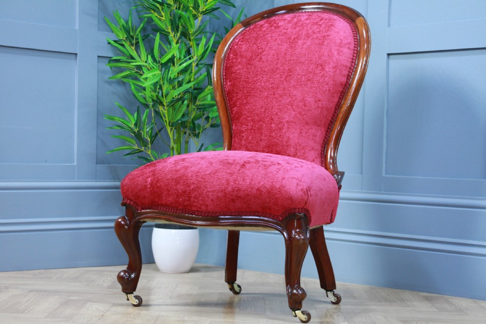 An antique Victorian balloon back nursing chair with a carved mahogany frame and deep pink velvet upholstery resting on castors. View II