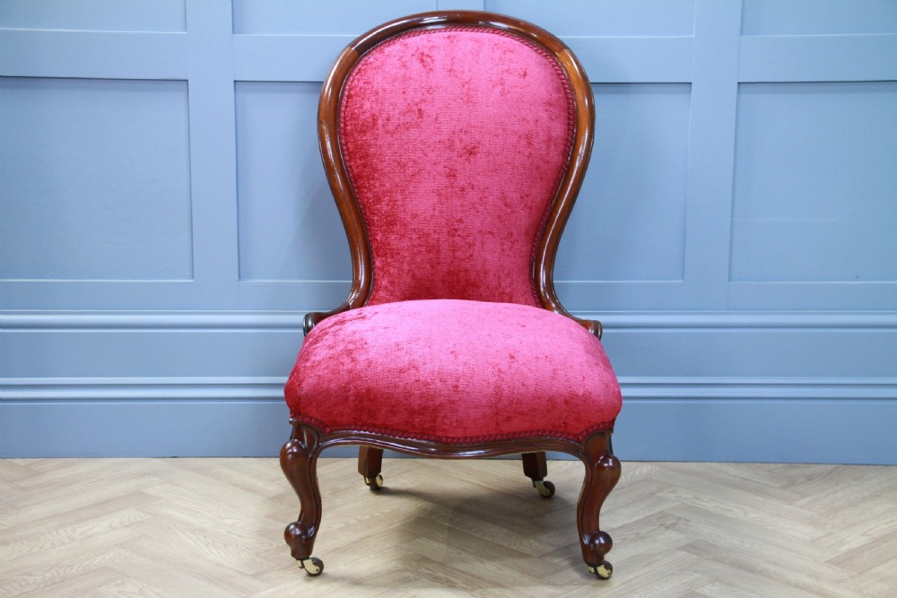 An antique Victorian balloon back nursing chair with a carved mahogany frame and deep pink velvet upholstery resting on castors. View III
