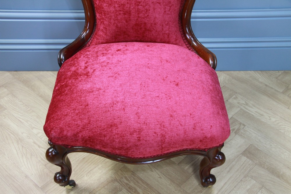 An antique Victorian balloon back nursing chair with a carved mahogany frame and deep pink velvet upholstery resting on castors. View IV