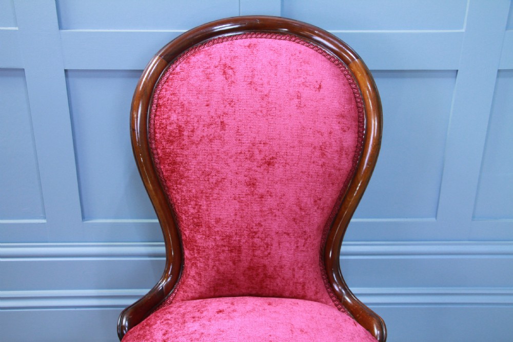 An antique Victorian balloon back nursing chair with a carved mahogany frame and deep pink velvet upholstery resting on castors. View V