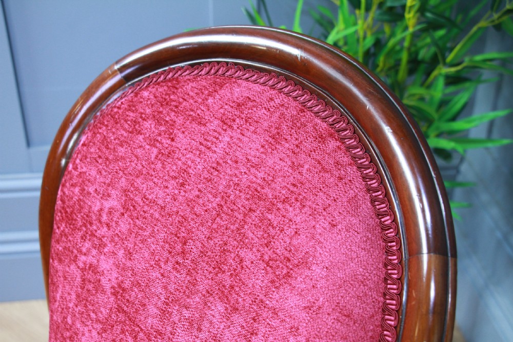 An antique Victorian balloon back nursing chair with a carved mahogany frame and deep pink velvet upholstery resting on castors. View VI