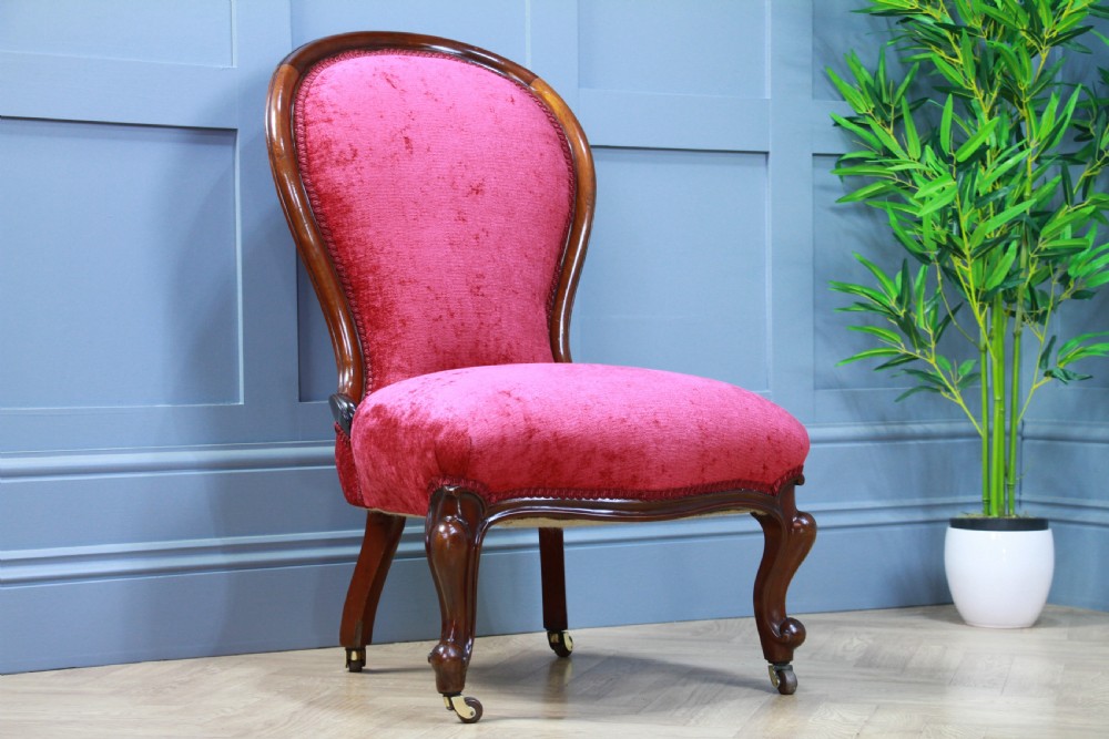 An antique Victorian balloon back nursing chair with a carved mahogany frame and deep pink velvet upholstery resting on castors. View X