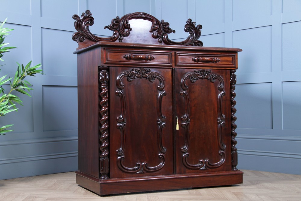A 19th-century dark mahogany chiffonier featuring two top drawers, two carved panel cupboard doors, barley twist side columns, and a carved scroll splashback. View X