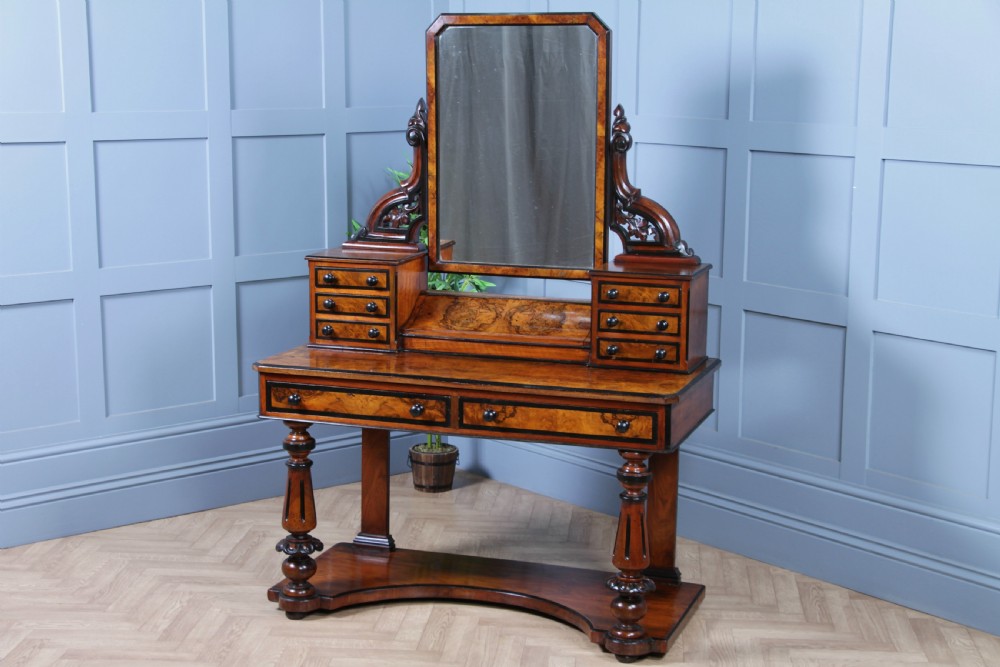 An antique Victorian burr walnut duchess dressing table featuring an integrated swing mirror, jewellery drawers, a curved piano top lid, turned front legs, and a plinth base.