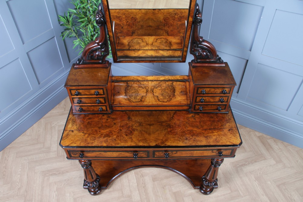 An antique Victorian burr walnut duchess dressing table featuring an integrated swing mirror, jewellery drawers, a curved piano top lid, turned front legs, and a plinth base. View V