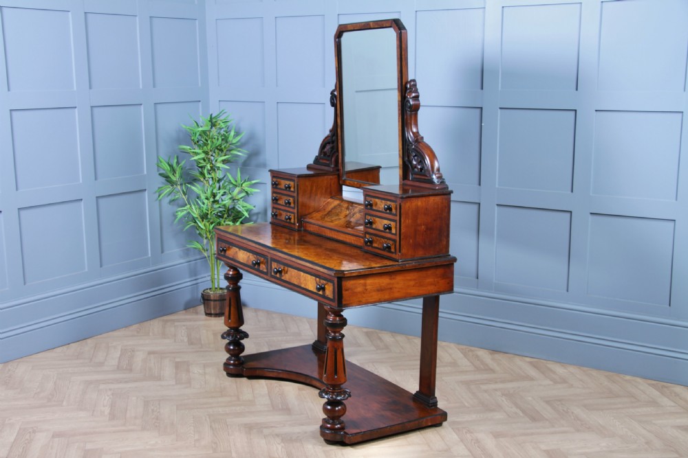 An antique Victorian burr walnut duchess dressing table featuring an integrated swing mirror, jewellery drawers, a curved piano top lid, turned front legs, and a plinth base. View XVI