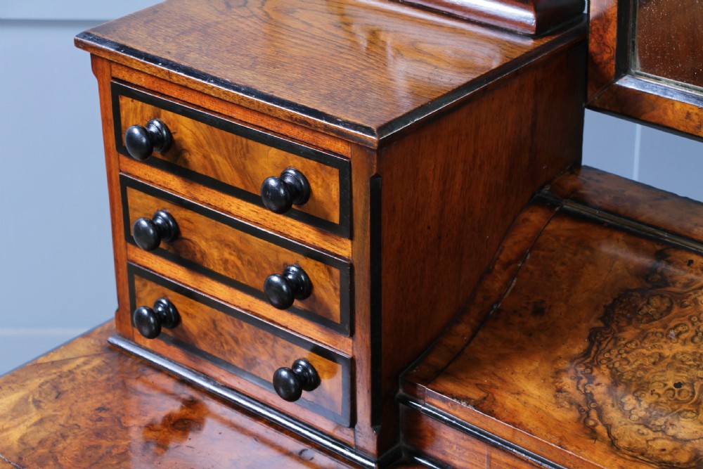 An antique Victorian burr walnut duchess dressing table featuring an integrated swing mirror, jewellery drawers, a curved piano top lid, turned front legs, and a plinth base. View XVIII
