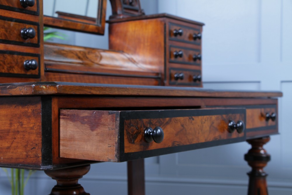 An antique Victorian burr walnut duchess dressing table featuring an integrated swing mirror, jewellery drawers, a curved piano top lid, turned front legs, and a plinth base. View XXI