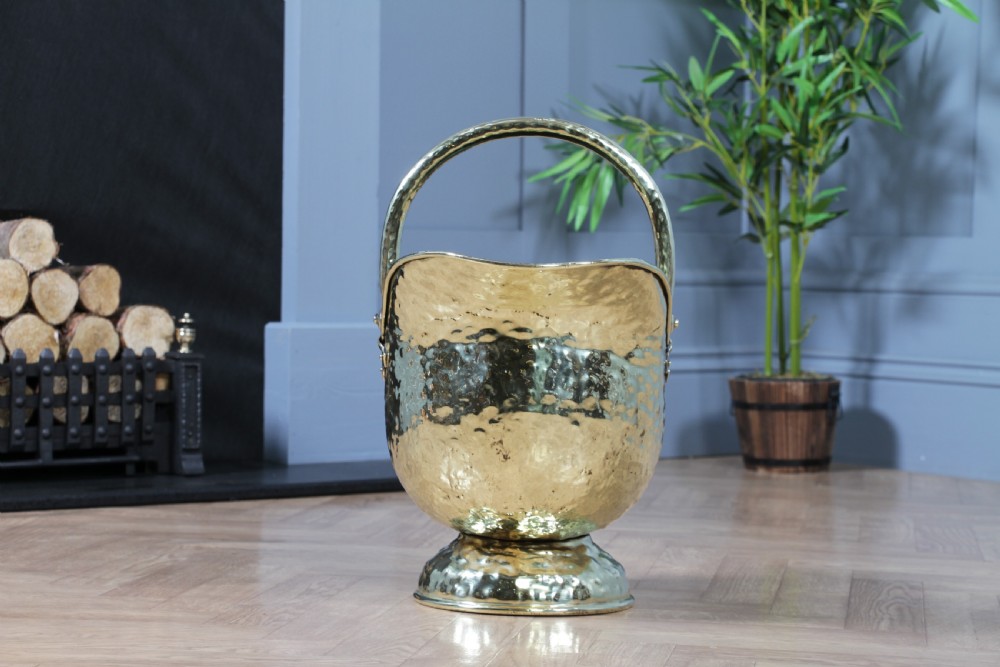 A Victorian hand-hammered brass helmet-shaped coal scuttle featuring a top swing handle, rear loop handle, and a round pedestal base. View V