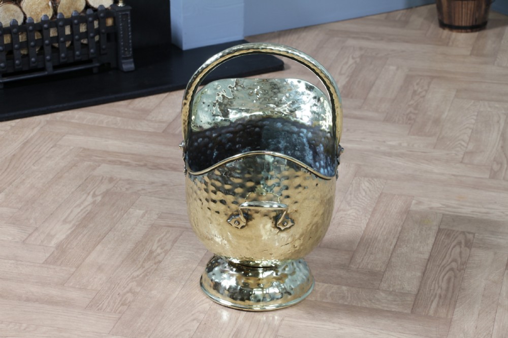 A Victorian hand-hammered brass helmet-shaped coal scuttle featuring a top swing handle, rear loop handle, and a round pedestal base. View VI