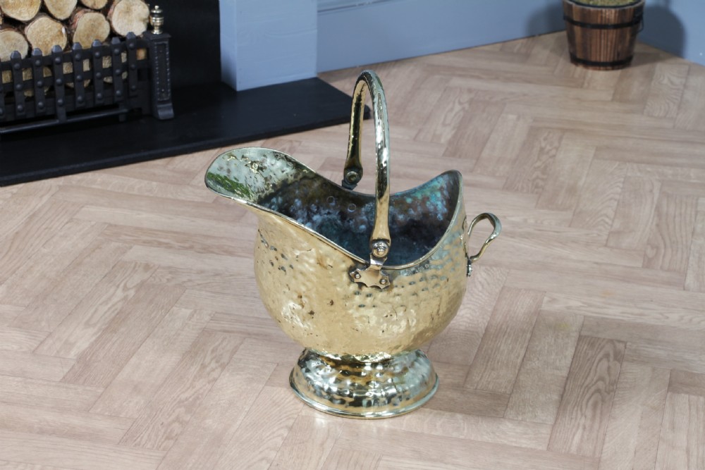 A Victorian hand-hammered brass helmet-shaped coal scuttle featuring a top swing handle, rear loop handle, and a round pedestal base. View VII