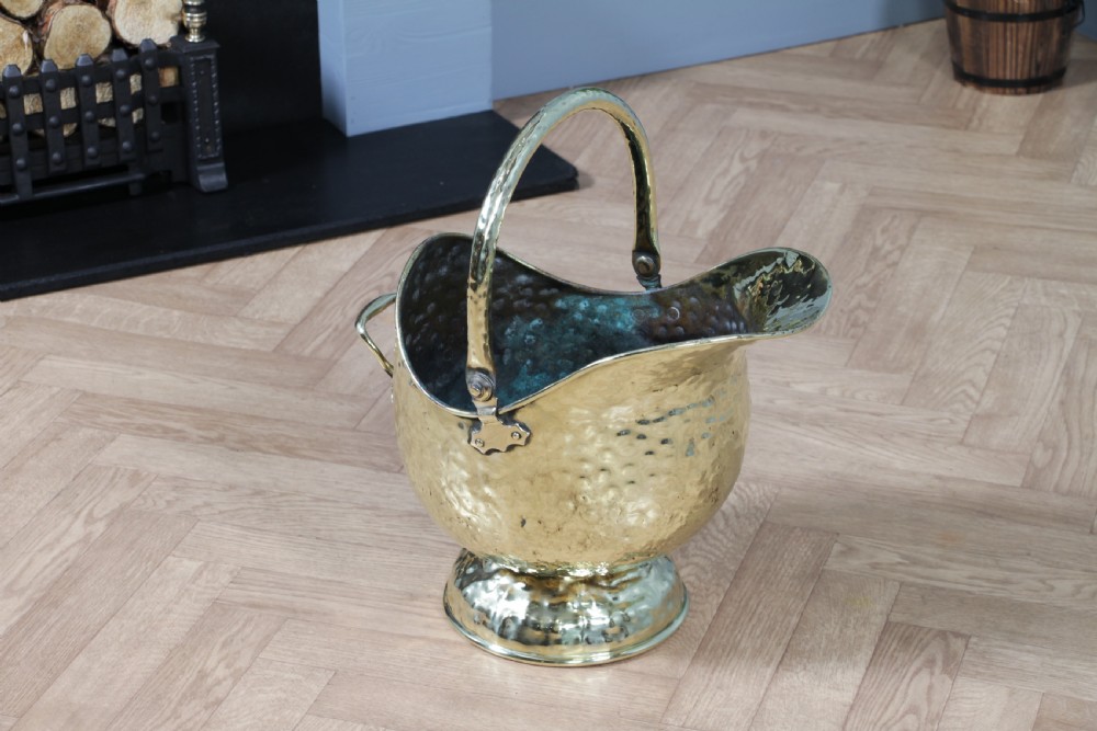 A Victorian hand-hammered brass helmet-shaped coal scuttle featuring a top swing handle, rear loop handle, and a round pedestal base. View VIII